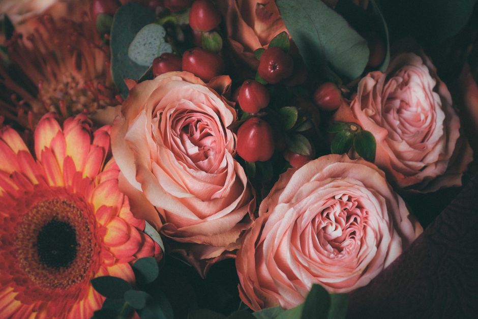 A close-up of a floral arrangement featuring three large, soft pink roses with delicate, layered petals and fresh green stems, complemented by bright orange and yellow gerbera daisies with dark center