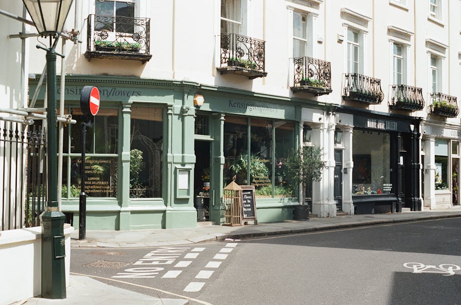 A storefront of a florist shop with a pale green facade, large glass windows displaying floral arrangements, and a black chalkboard sign outside. The shop is situated on a street corner with a pedestr