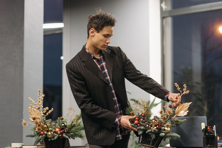 A young man in a dark pinstripe blazer and plaid shirt arranges a festive floral centerpiece with a variety of greenery, including pine branches, and decorative elements such as red, gold, and bronze