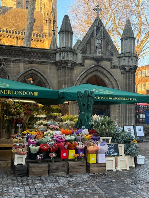 A vibrant outdoor flower stall situated in front of a historic stone building with Gothic architectural elements, including pointed arches and decorative pinnacles. The stall, managed by flowersholbor